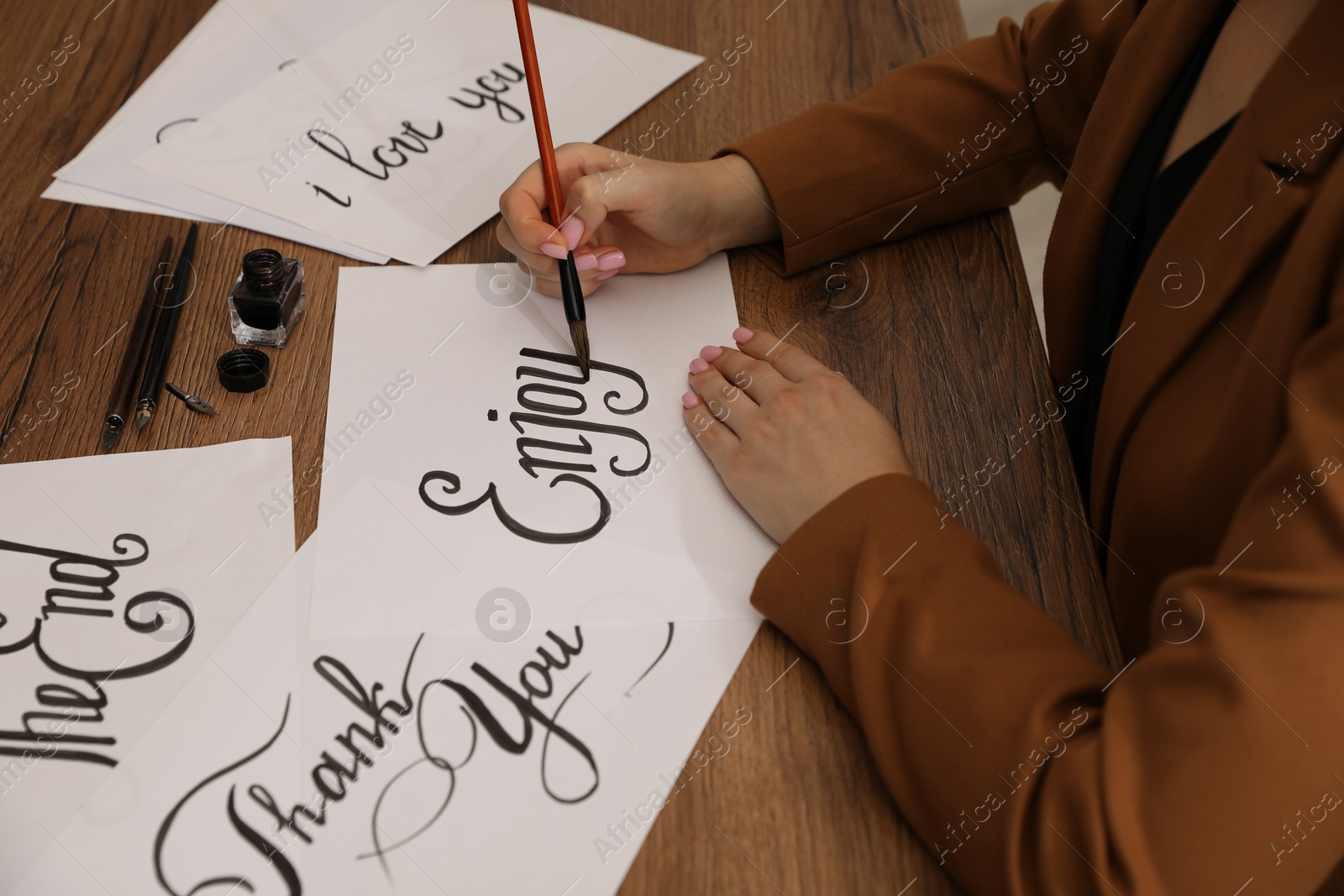 Calligraphy. Woman with brush writing word Enjoy on sheet of paper at wooden table, closeup Photo of Calligraphy. Woman with brush writing word Enjoy on sheet of paper at wooden table, closeup