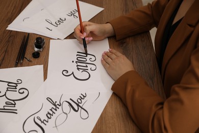 Photo of Calligraphy. Woman with brush writing word Enjoy on sheet of paper at wooden table, closeup