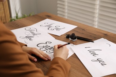 Photo of Calligraphy. Woman with brush writing word Enjoy on sheet of paper at wooden table, closeup