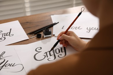 Photo of Calligraphy. Woman with brush writing word Enjoy on sheet of paper at wooden table, closeup