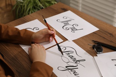 Photo of Calligraphy. Woman with brush writing words Thank You on sheet of paper at wooden table, closeup