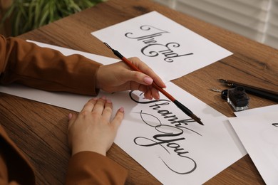 Photo of Calligraphy. Woman with brush writing words Thank You on sheet of paper at wooden table, closeup