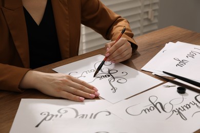 Photo of Calligraphy. Woman with brush writing phrase I Love You on sheet of paper at wooden table, closeup