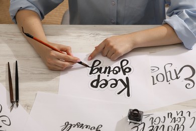 Photo of Calligraphy. Woman with brush writing phrase Keep Going on sheet of paper at wooden table, closeup