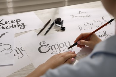 Photo of Calligraphy. Woman with brush writing word Enjoy on sheet of paper at wooden table, closeup