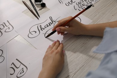 Photo of Calligraphy. Woman with brush writing words Thank You on sheet of paper at wooden table, above view