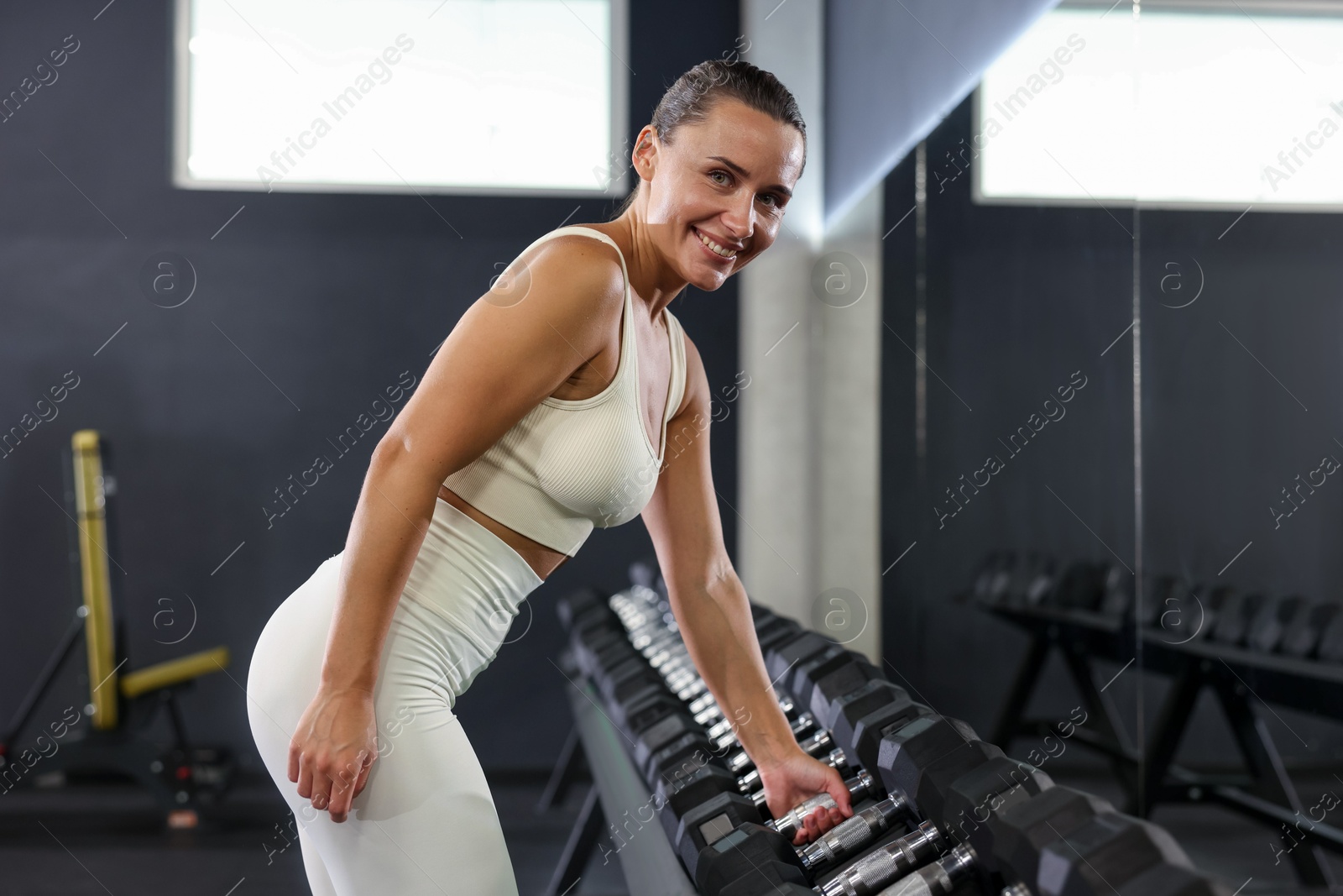 Photo of Smiling woman taking dumbbell near mirror in gym