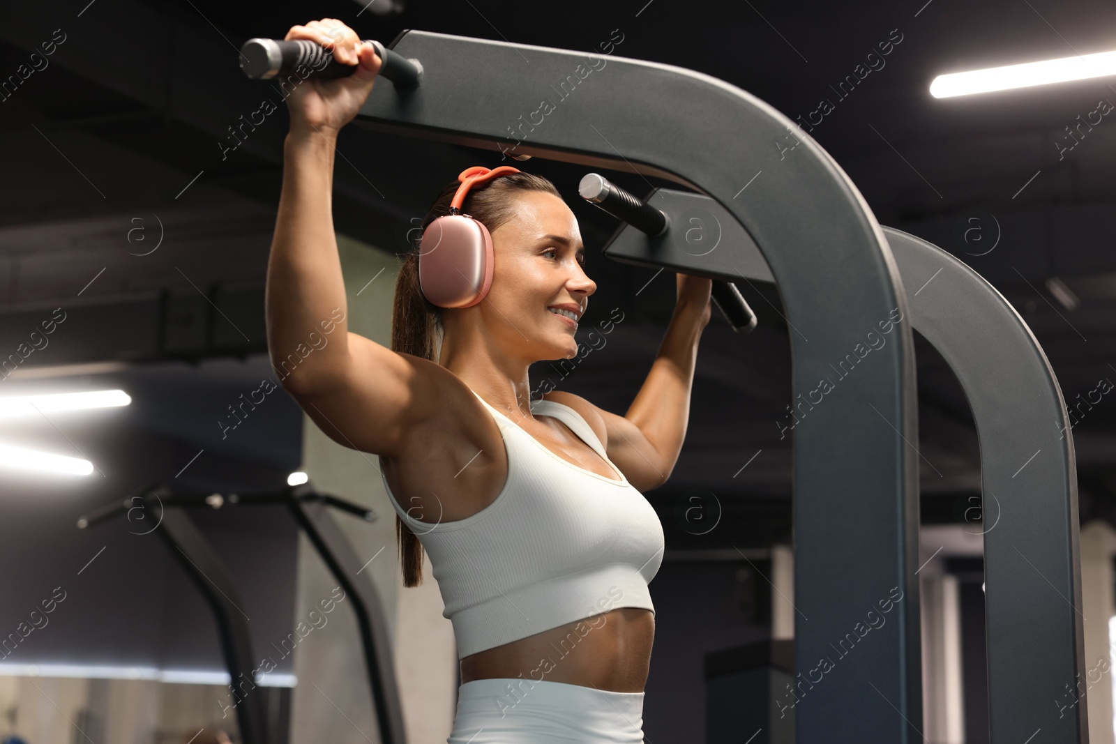 Photo of Smiling woman in headphones training on pull up machine in gym