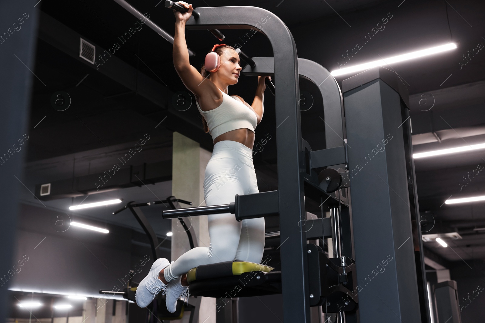 Photo of Athletic woman in headphones training on pull up machine in gym, low angle view