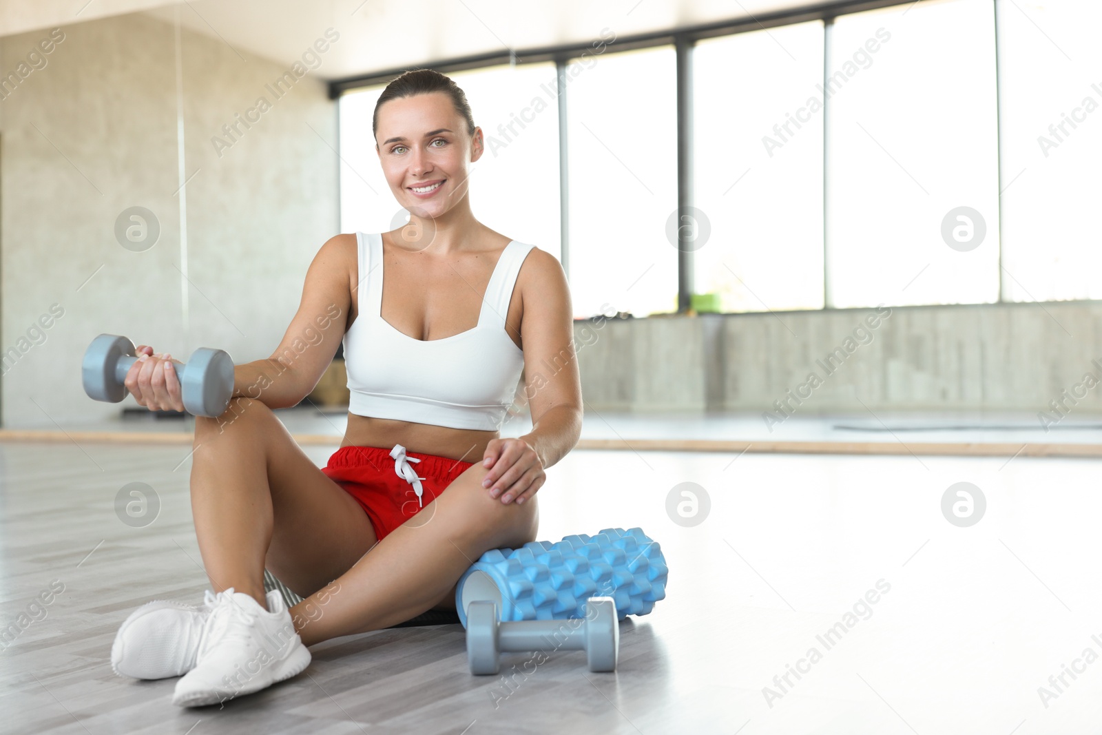 Smiling woman with dumbbells and foam roller on floor in gym. Space for text Photo of Smiling woman with dumbbells and foam roller on floor in gym. Space for text