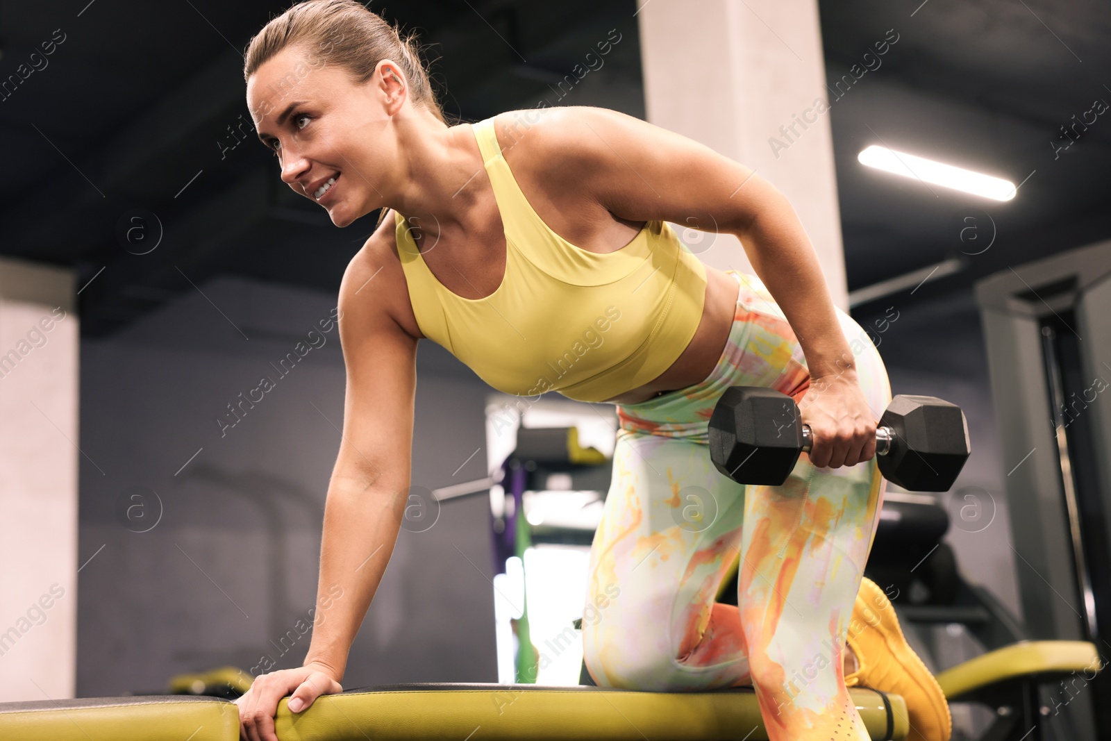 Photo of Smiling woman training with dumbbell on exercise bench in gym, low angle view