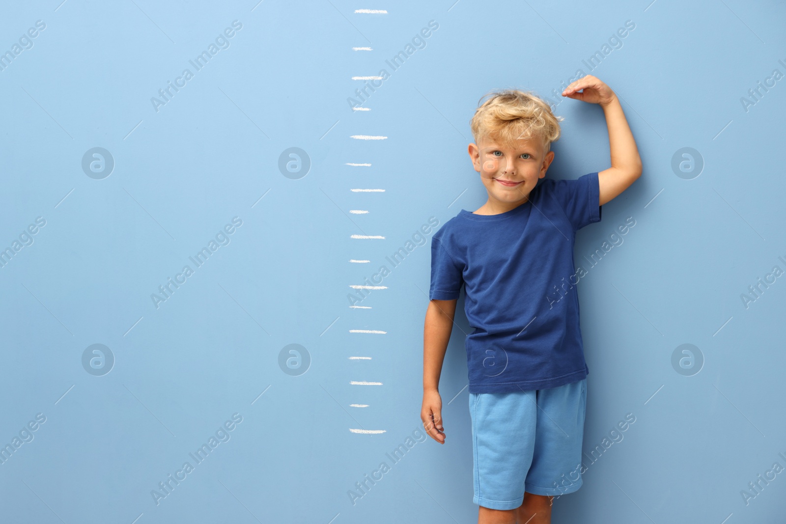 Photo of Cute little boy measuring his height near blue wall. Space for text