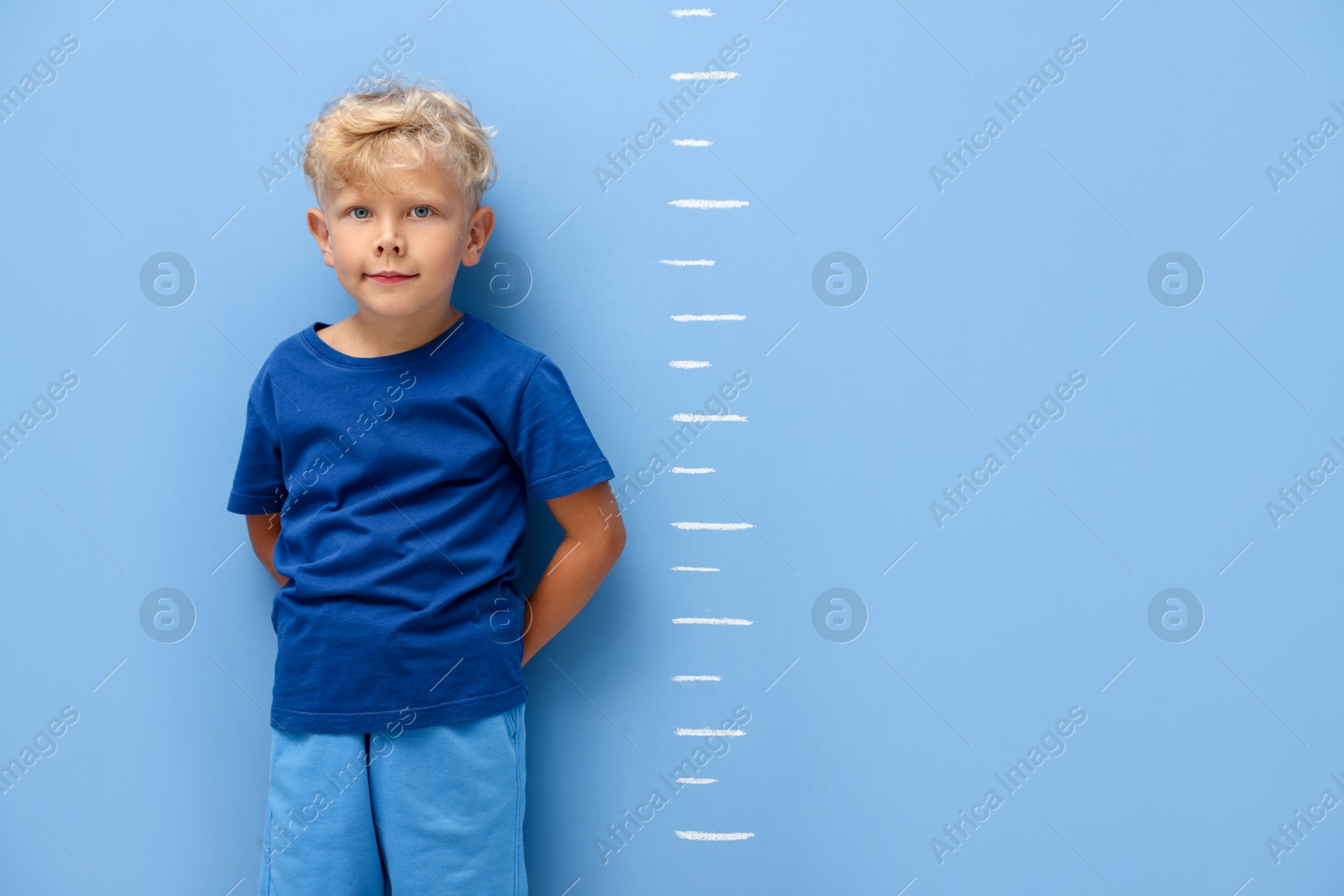 Photo of Cute little boy measuring his height near blue wall. Space for text