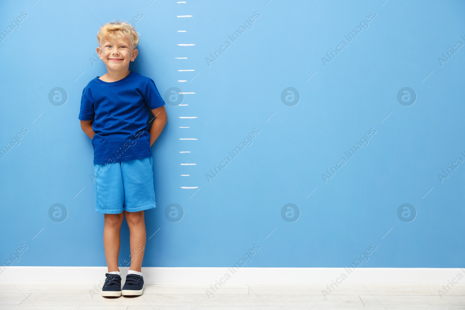 Photo of Cute little boy measuring his height near blue wall. Space for text