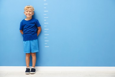Photo of Cute little boy measuring his height near blue wall. Space for text