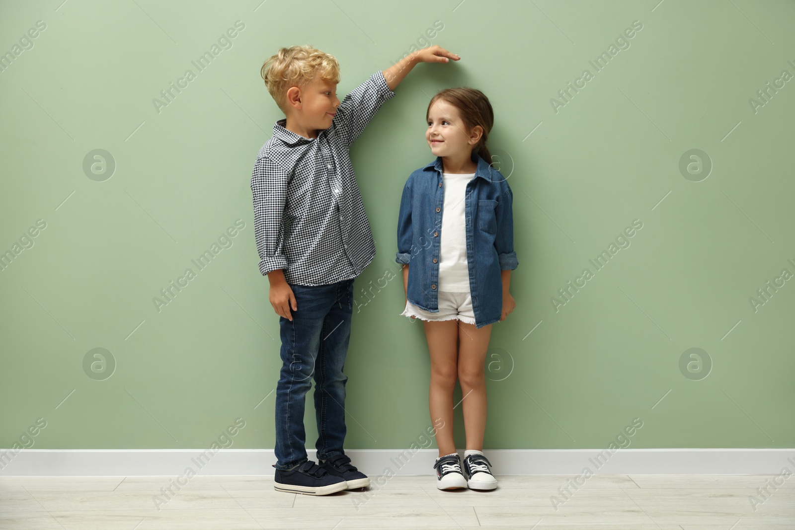 Photo of Cute little girl and boy measuring their heights near green wall