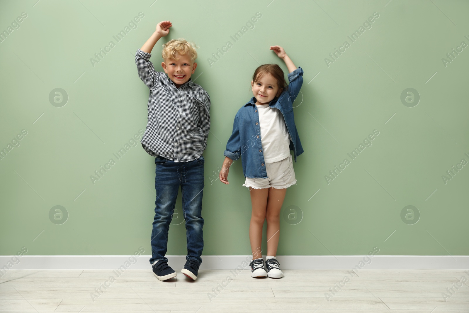 Photo of Cute little girl and boy measuring their heights near green wall