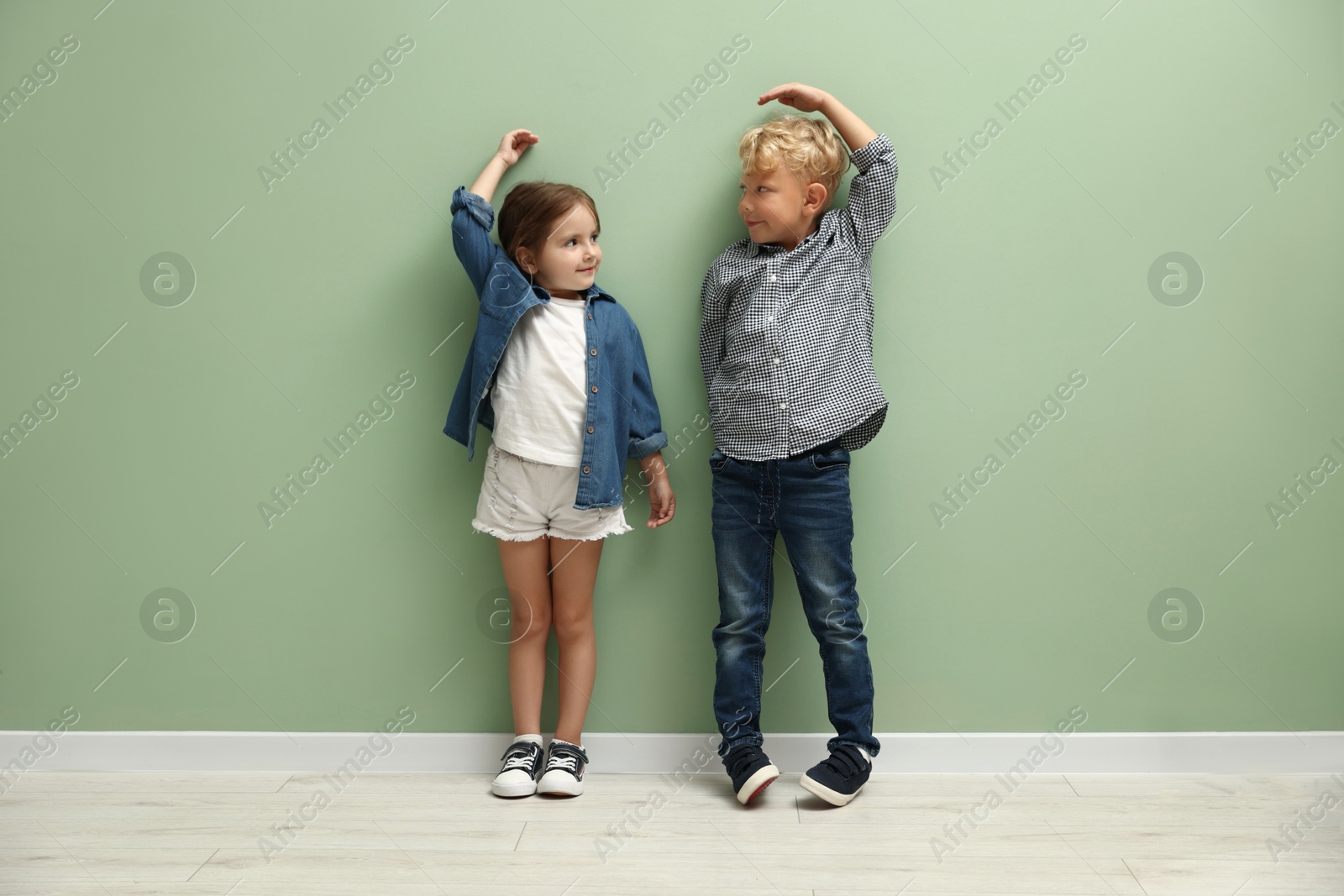 Photo of Cute little girl and boy measuring their heights near green wall