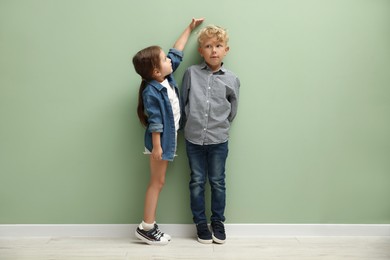 Photo of Cute little girl and boy measuring their heights near green wall