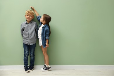 Photo of Cute little girl and boy measuring their heights near green wall. Space for text