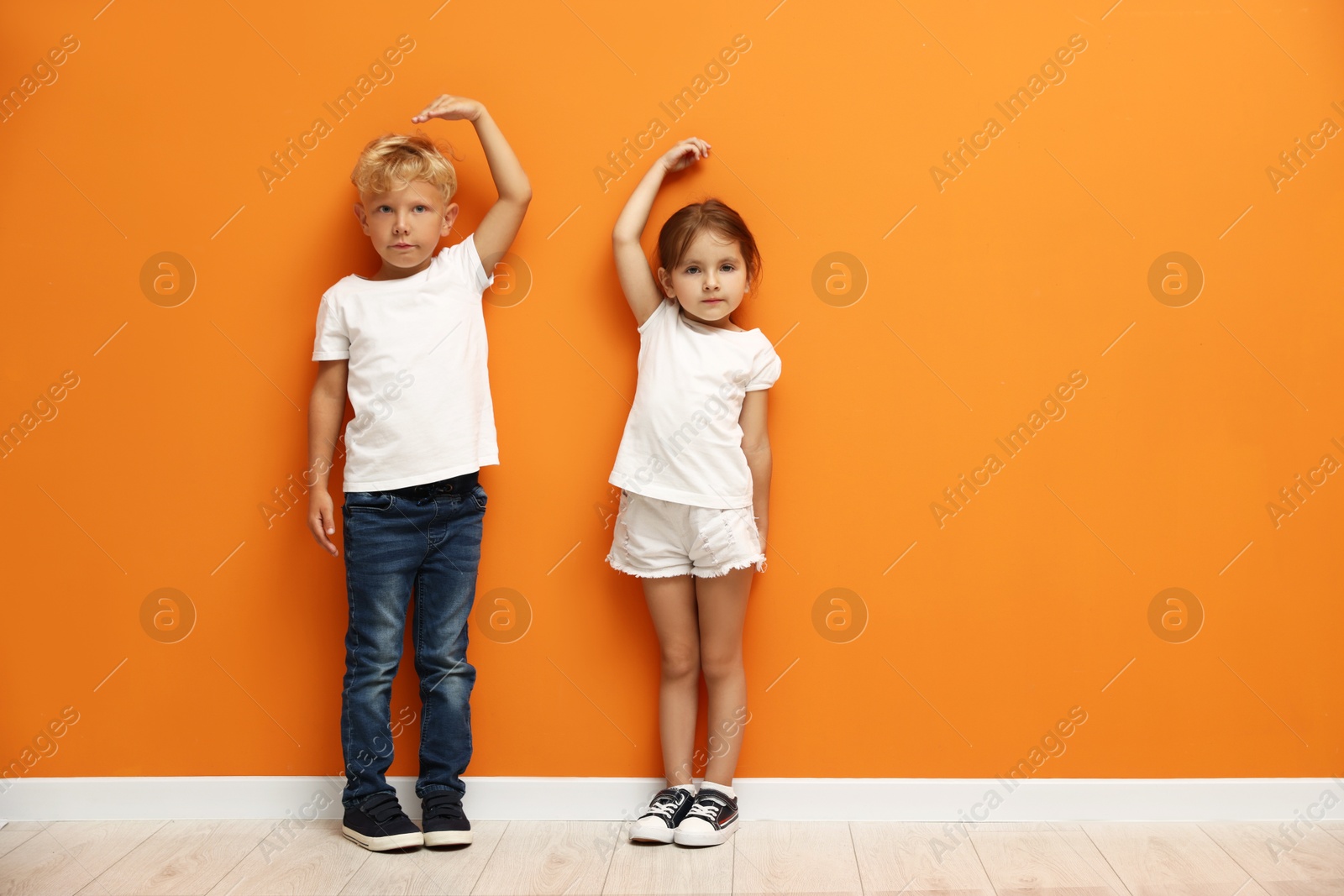 Photo of Cute little girl and boy measuring their heights near orange wall