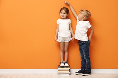 Photo of Cute little girl and boy measuring their heights near orange wall. Space for text
