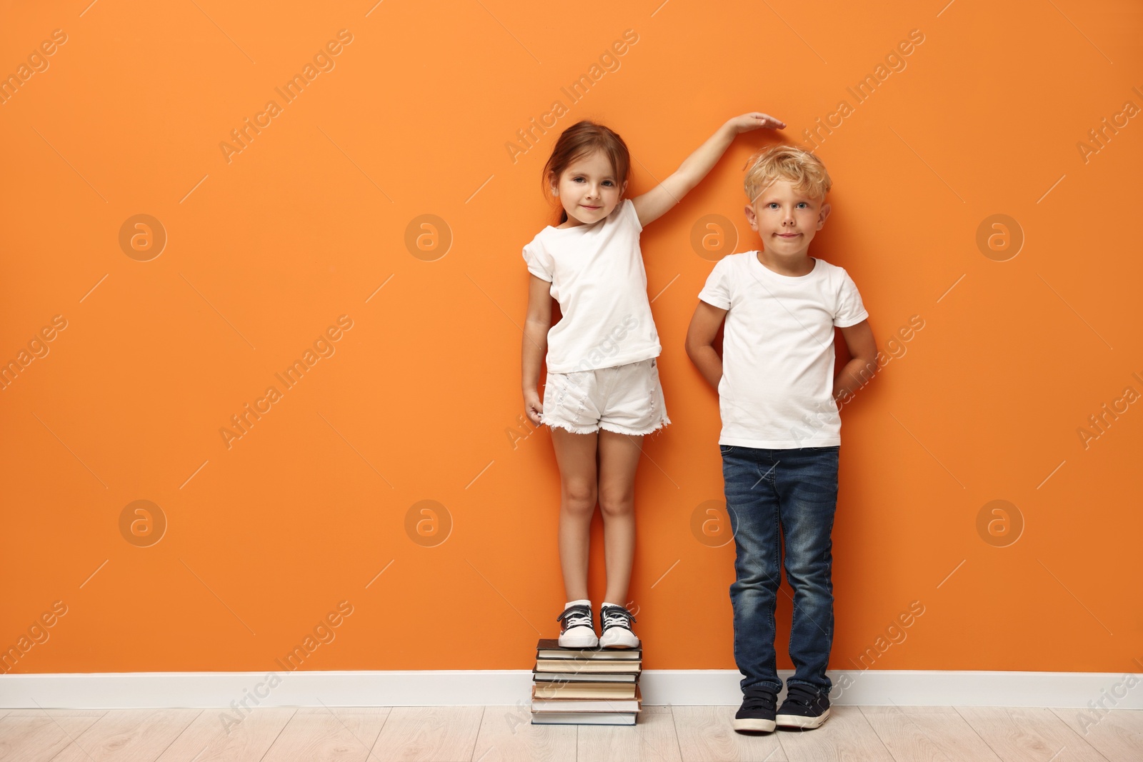 Photo of Cute little girl and boy measuring their heights near orange wall. Space for text