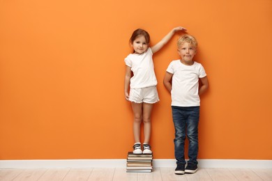 Photo of Cute little girl and boy measuring their heights near orange wall. Space for text