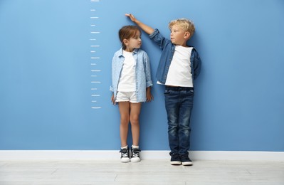 Photo of Cute little girl and boy measuring their heights near blue wall