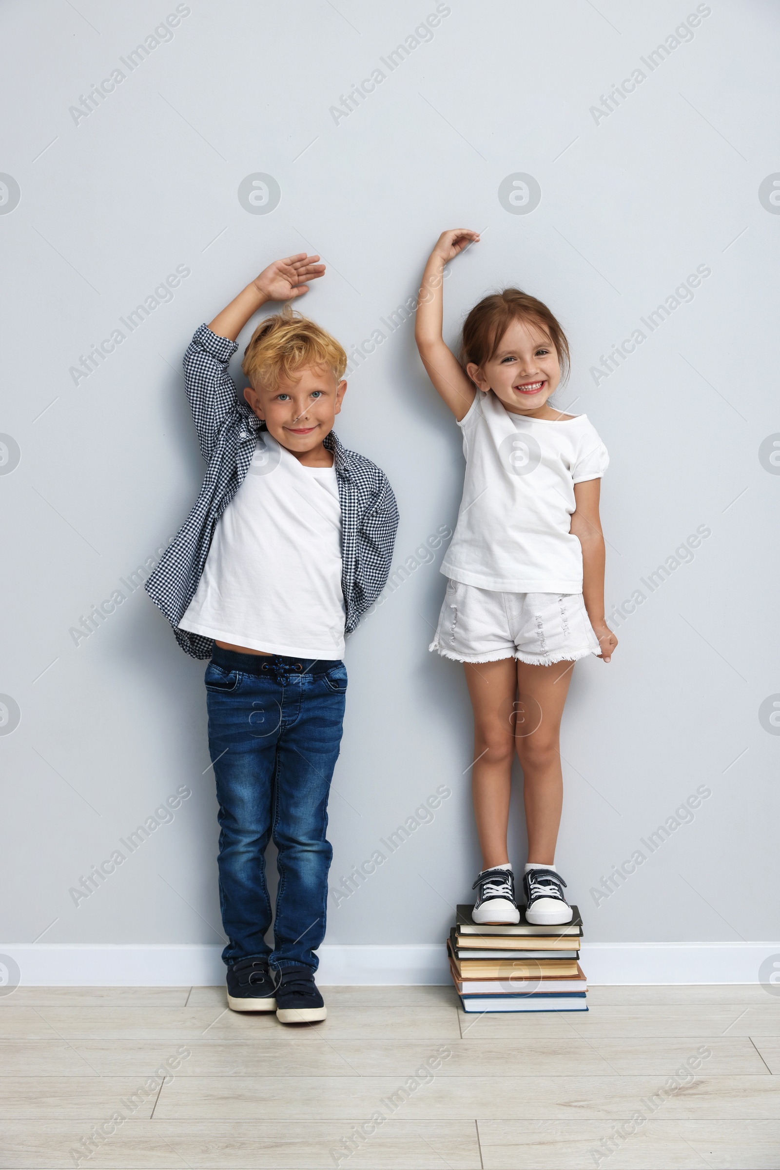 Photo of Cute little girl and boy measuring their heights near light wall