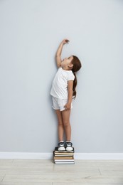 Photo of Cute little girl on stack of books measuring her height near light wall