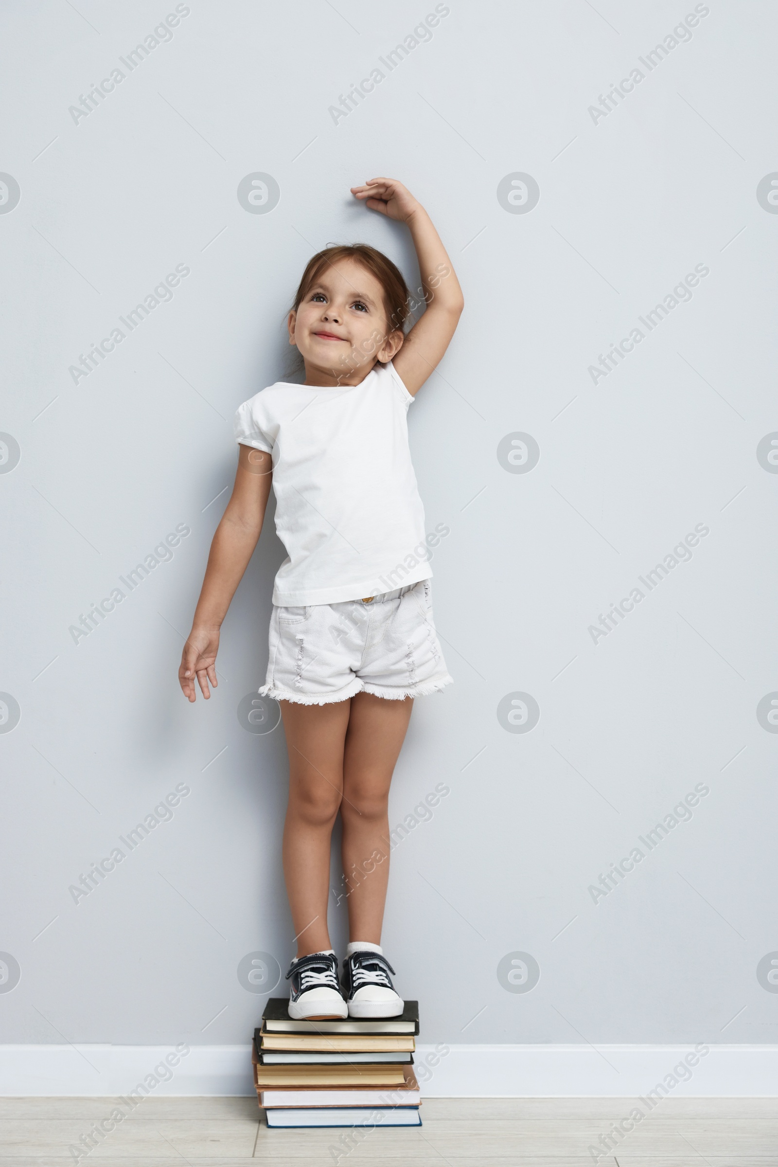 Photo of Cute little girl on stack of books measuring her height near light wall