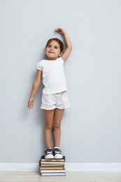 Photo of Cute little girl on stack of books measuring her height near light wall