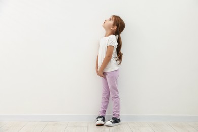 Photo of Cute little girl standing near white wall and looking up. Space for text