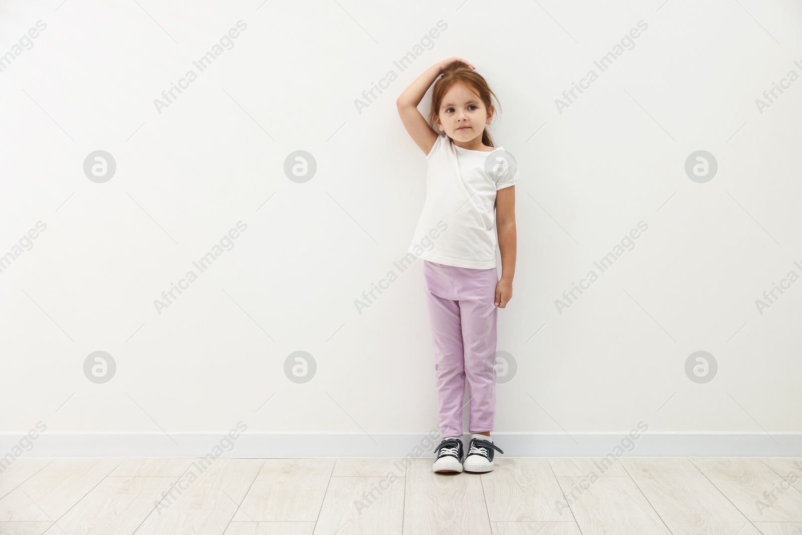 Photo of Cute little girl measuring her height near white wall. Space for text