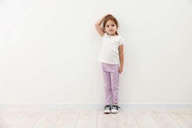 Photo of Cute little girl measuring her height near white wall. Space for text