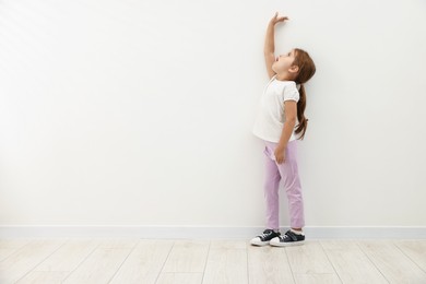 Photo of Cute little girl measuring her height near white wall. Space for text