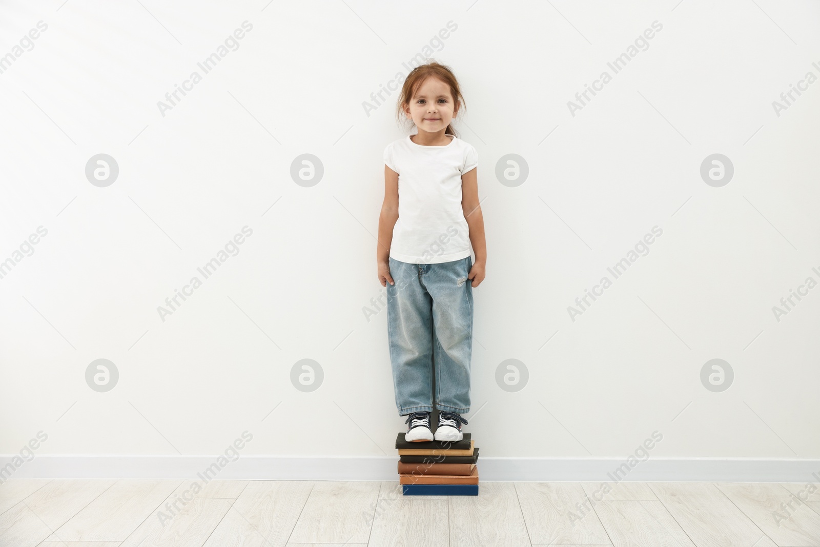 Photo of Cute little girl on stack of books measuring her height near white wall