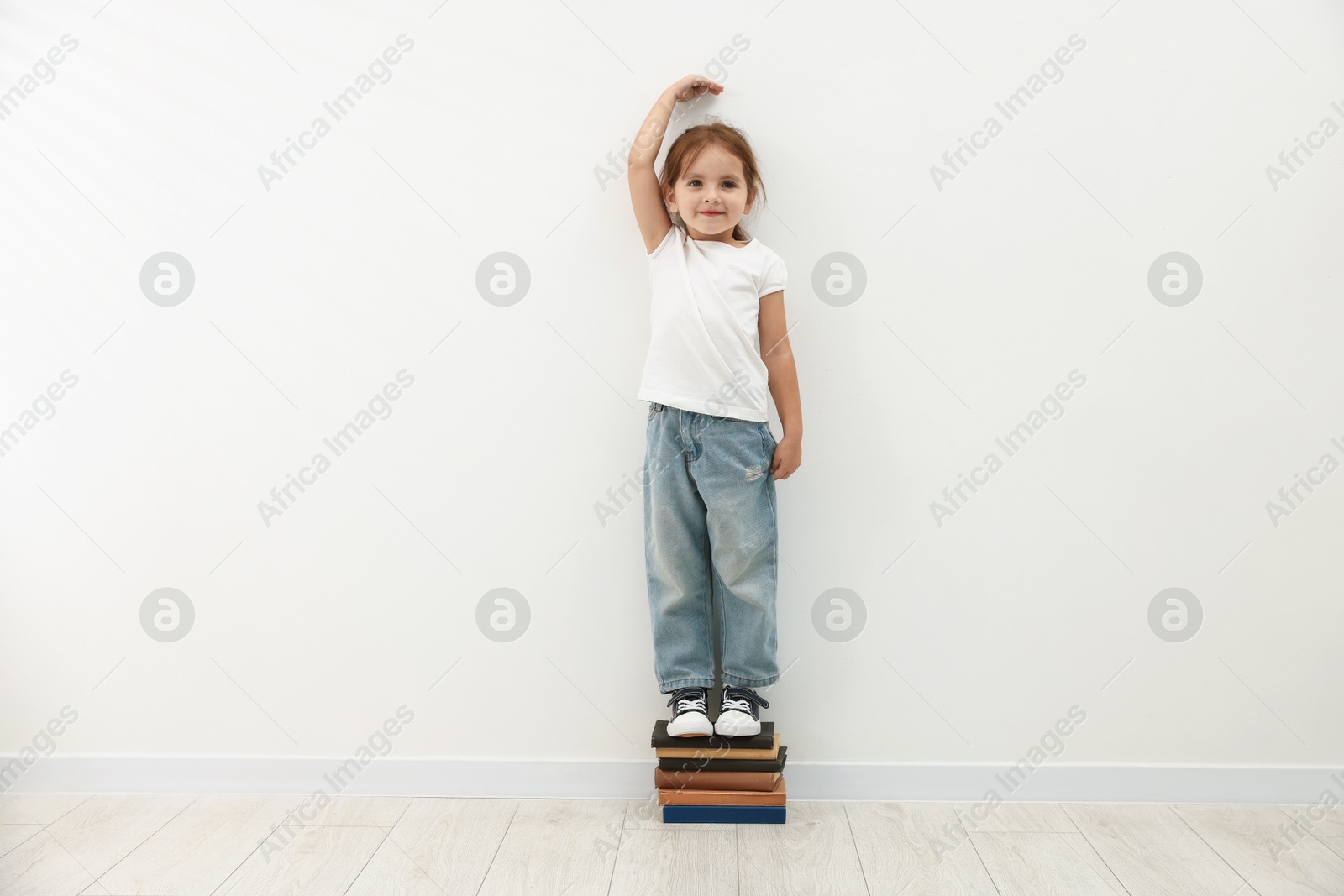 Photo of Cute little girl on stack of books measuring her height near white wall