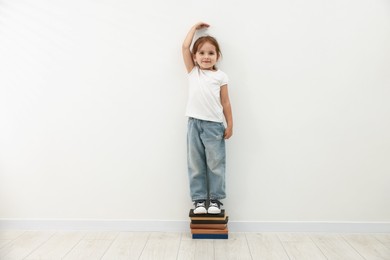 Photo of Cute little girl on stack of books measuring her height near white wall