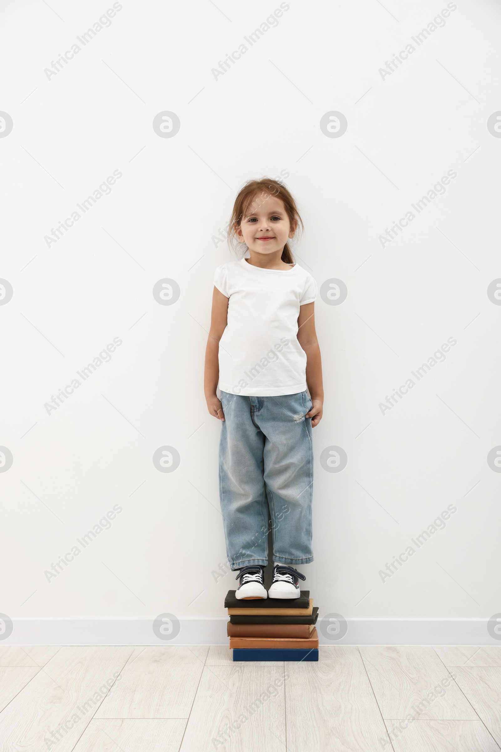 Photo of Cute little girl on stack of books measuring her height near white wall