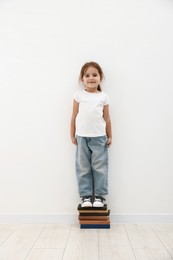 Photo of Cute little girl on stack of books measuring her height near white wall