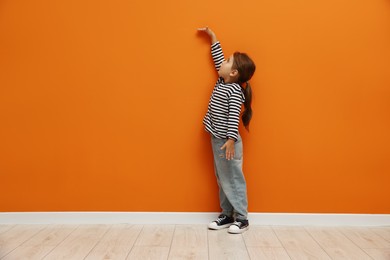Photo of Cute little girl measuring her height near orange wall. Space for text