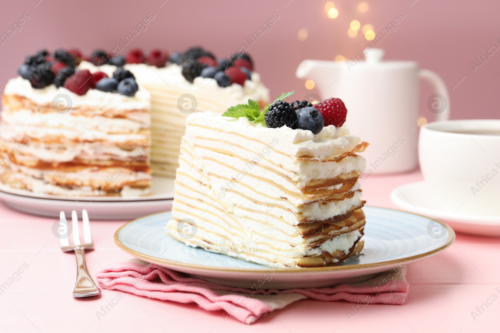 Photo of Piece of tasty crepe cake with berries served on pink wooden table against blurred lights, closeup