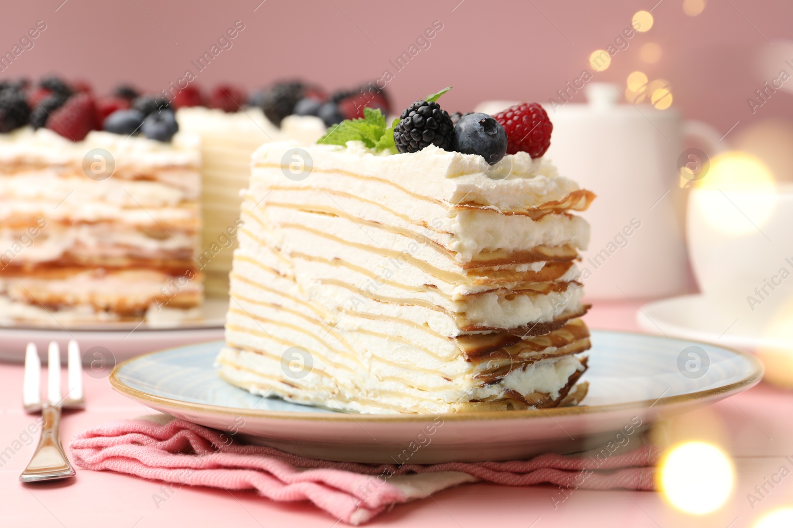 Photo of Piece of tasty crepe cake with berries served on pink wooden table against blurred lights, closeup