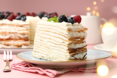 Photo of Piece of tasty crepe cake with berries served on pink wooden table against blurred lights, closeup