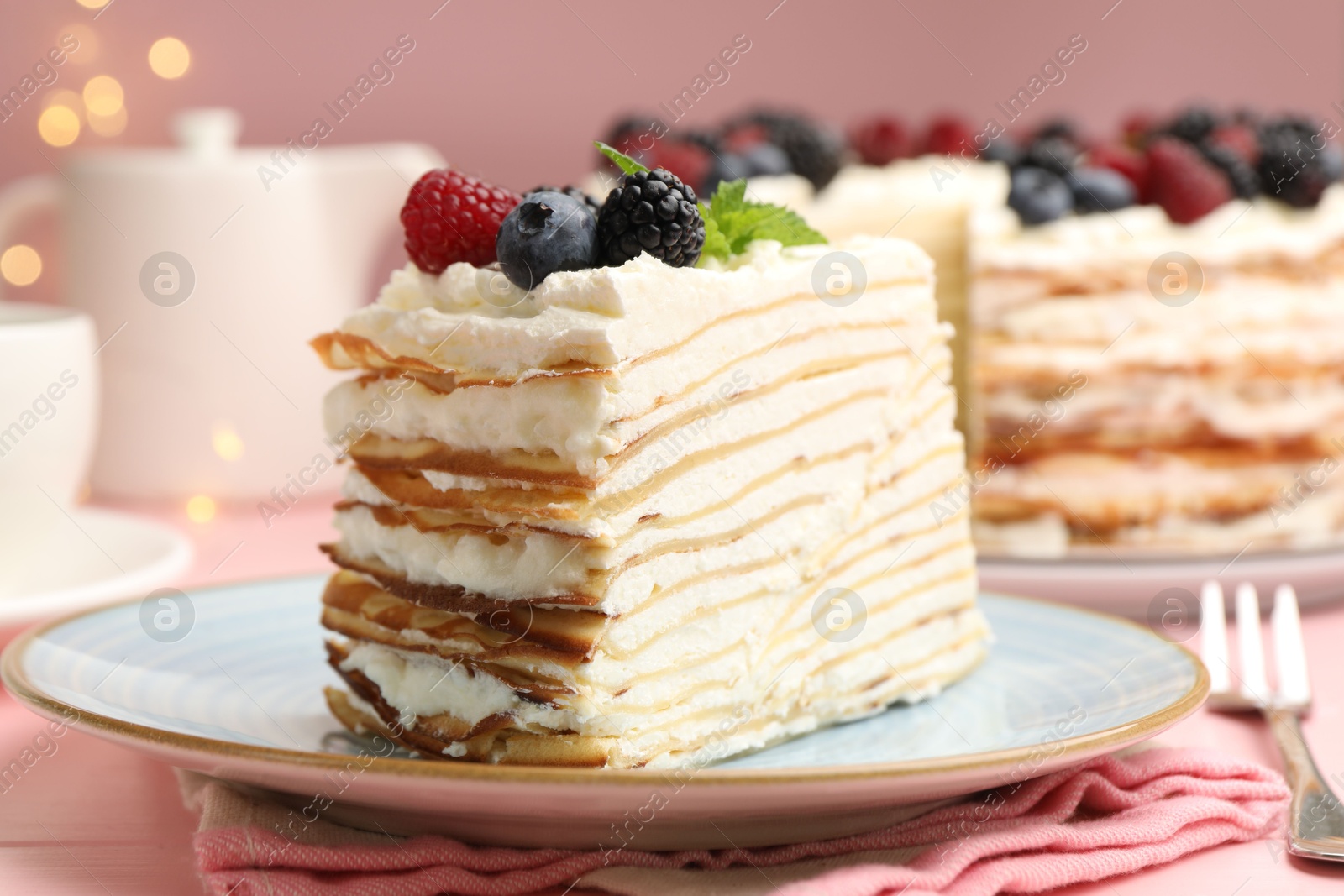 Photo of Piece of tasty crepe cake with berries served on pink wooden table against blurred lights, closeup