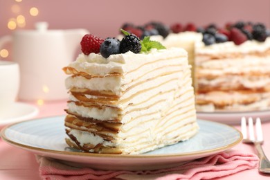 Photo of Piece of tasty crepe cake with berries served on pink wooden table against blurred lights, closeup