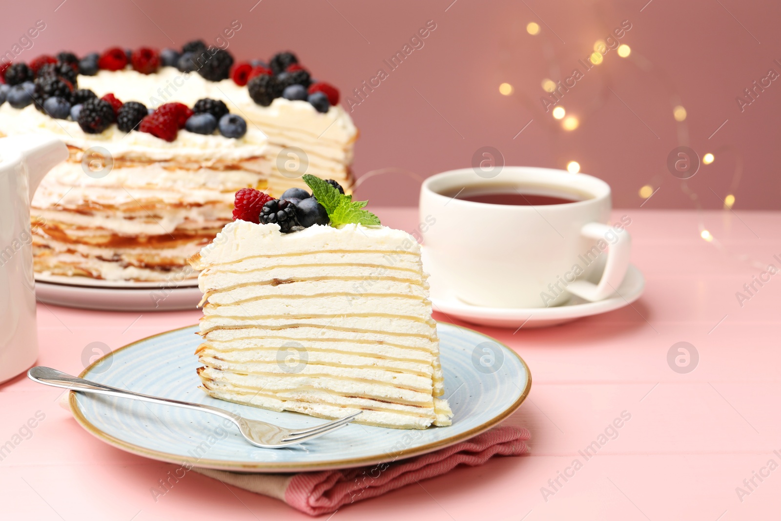 Photo of Piece of tasty crepe cake with berries served on pink wooden table against blurred lights, closeup