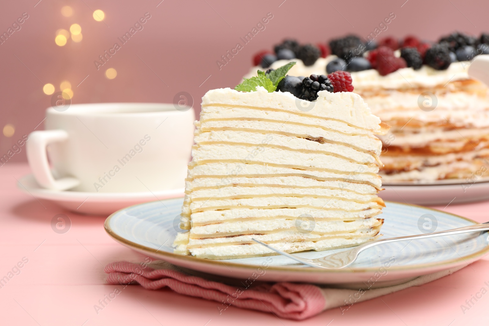 Photo of Piece of tasty crepe cake with berries served on pink wooden table against blurred lights, closeup